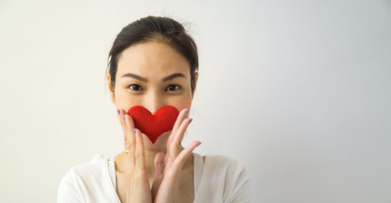 Eine Person in einem weißen Hemd hält ein Herz aus FIlz vor ihren Mund. 