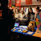 Personen in einem Marktstand, auf der Tischfläche Flyer und ein kleines Glücksrad