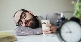 Dunkelhaariger Mann mit schwarzer Hornbrille schläft an einem weißen Tisch. Er hält einen Kaffeebecher mit der Aufschrift "Coffee". Auf dem Tisch befinden sich zusätzlich eine Uhr und ein Buch (beide verschwommen)