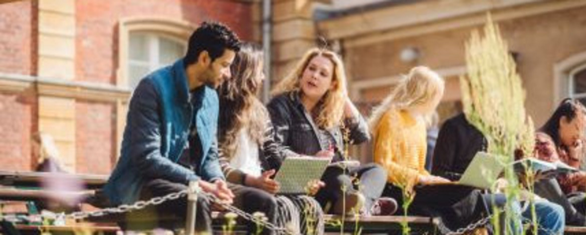 Studierende sitzen auf einer Bank am Campus Neues Palais und unterhalten sich. Eine Studentin hat einen Laptop auf dem Schoß.