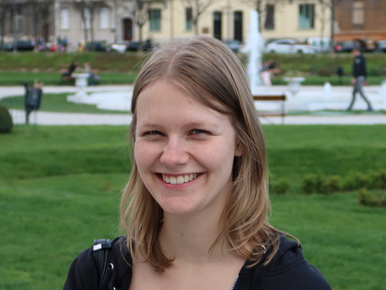 A portrait of Vivien Fröhlich. She is a young woman with pale skin and shoulder length dark blonde hair. She is wearing a teal shirt and black hoodie. She is standing in a park.