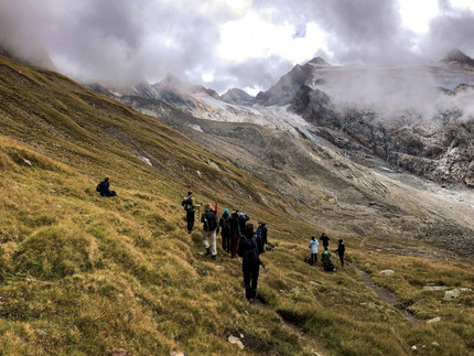 Studierende stehen auf einer Wiese, welchen auf einen steinigen Berg hinausläuft. Tief über den Studierenden hängen Wolken.