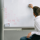 Ein Mann sitzt im Rollstuhl, er trägt ein weißes Hemd und zeichnet etwas auf einem Whiteboard an. 