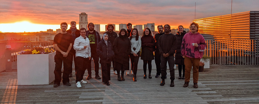 Eine Gruppe junger Menschen steht auf einer Dachterrasse und lächelt in die Kamera. Im Hintergrund sieht man die Dächer Potsdams und einen Sonnenuntergang.