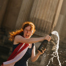 Ein*e lachende Absolvent*in mit rotem Schal hält eine Flasche aus der Champagner sprudelt