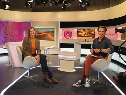 Prof. Linstädter and journalist sitting in a recording studio with cameras and screens with pictures of the desert in the background.