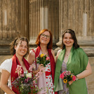 Drei Absolvent*innen posieren mit Blumensträßen vor den Säulen des Kolonnadenbogens.