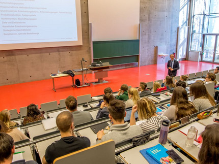 Ein Mann steht in einem Hörsaal vor vielen Studierenden und hält eine Vorlesung