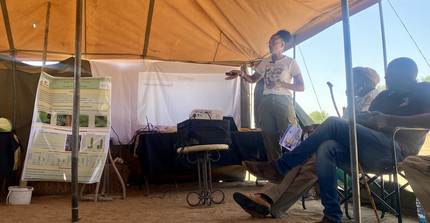 Researchers standing in front of a poster under a tent talking to a small audience of seated people