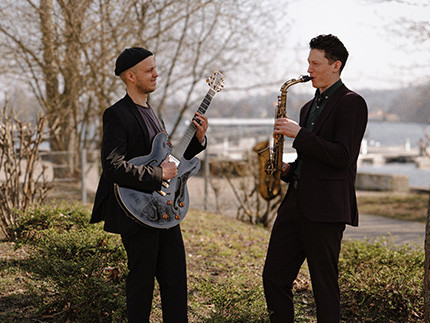 Ein Mann mit Gitarre und ein Mann mit Saxophon stehen auf einer Wiese, im Hintergrund sieht man Wasser und Boote