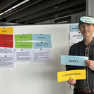 Eine männlich gelesene Person hält vor einer Moderationswand drei Moderationskarten hoch.