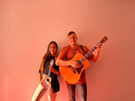 Zwei Personen mit Gitarren in der Hand vor einer orangenen Wand