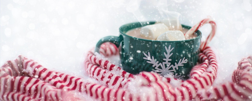 Grüne Tasse mit heißem Kakao und Marshmallows steht im Schnee und ist umgeben von einem weiß-roten Schal.