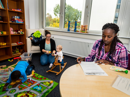 woman plays with child in a room, an other child is playing on the floor and a second woman is sitting on a chair and makes some notes