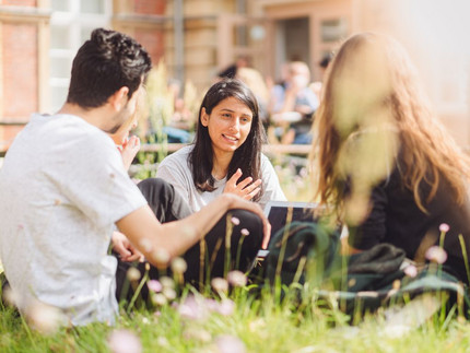 Drei Studierende sitzen auf dem Campus Neues Palais auf einer Wiese und unterhalten sich.