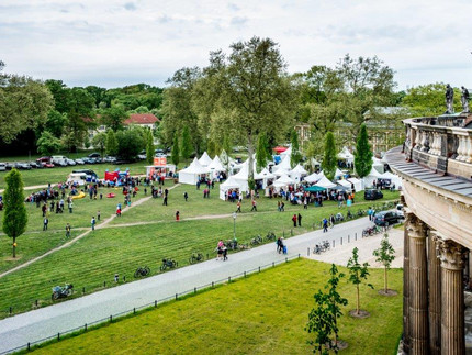 Die Grasfläche vor den Kolonaden der Universität Potsdam fotografiert von hoch oben. Im Hintergrund auf dem Rasen sind weiße Zelte aufgestellt. Im Vordergrund der rechten Seite ist ein Teil der Kolonaden sichtbar. Das Gras ist Grün und der Himmel leicht bewölkt. Menschen halten sich auf dem Rasen auf.