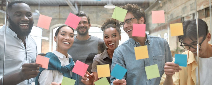Verschiedene, junge Menschen stehen vor einer Glaswand, die mit bunten Klebezetteln beklebt ist.