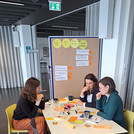Verschiedene Menschen sitzen an Tischen im Rahmen eines Workshopsettings und haben viele verschiedene Materialien auf dem Tisch.