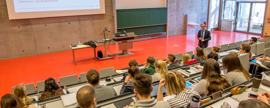 Ein Mann steht in einem Hörsaal vor vielen Studierenden und hält eine Vorlesung