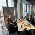 Verschiedene Menschen sitzen an Tischen im Rahmen eines Workshopsettings und haben viele verschiedene Materialien auf dem Tisch.