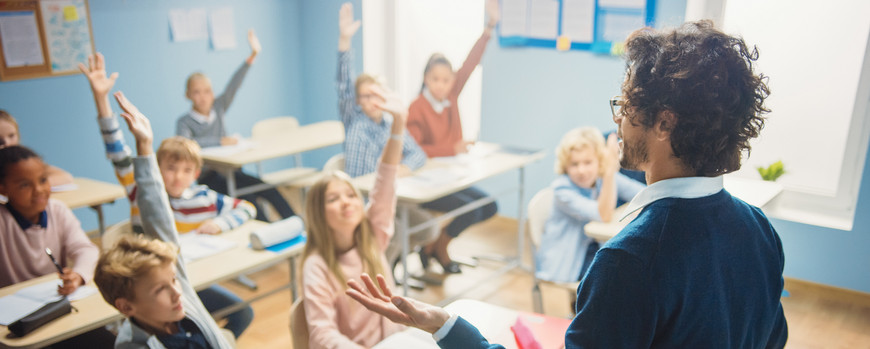 Lehrer steht vor einer Klasse und fragt die Schüler etwas. Diese melden sich begeistert.