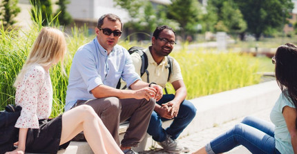 Studierende am Campus Golm der Universität Potsdam. Studierende sitzen auf dem Campus Golm auf einer Bordsteinkante. Hinter ihnen ist eine Wiese zu sehen.